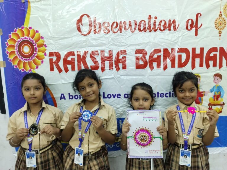 Four young girls in beige school uniforms with lanyards stand in front of a Rakshabandhan banner, proudly displaying handmade bracelets and crafts.